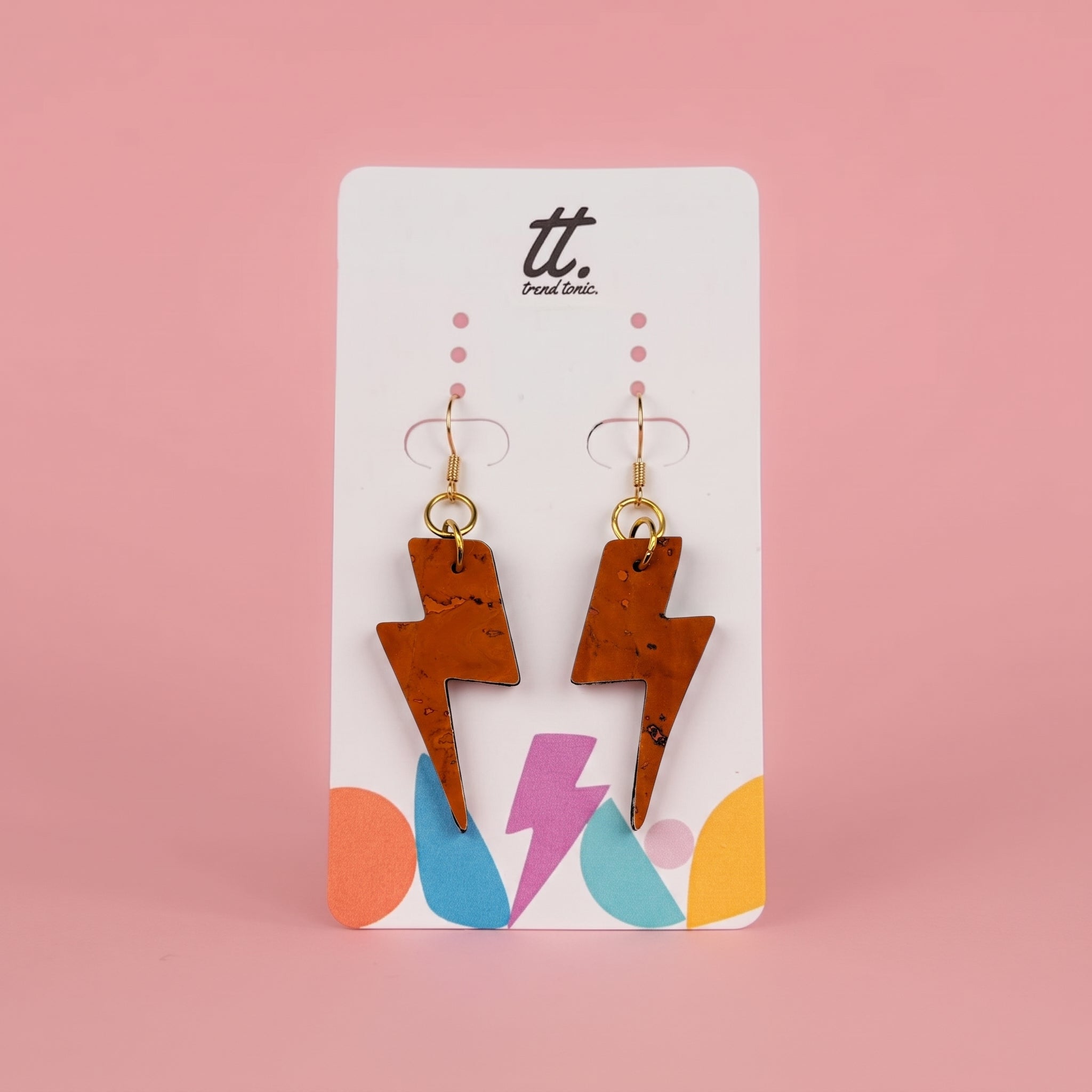 A pair of cork lightning bolt drop earrings with gold hooks, displayed on a white brand card with colorful accents against a solid pink background.