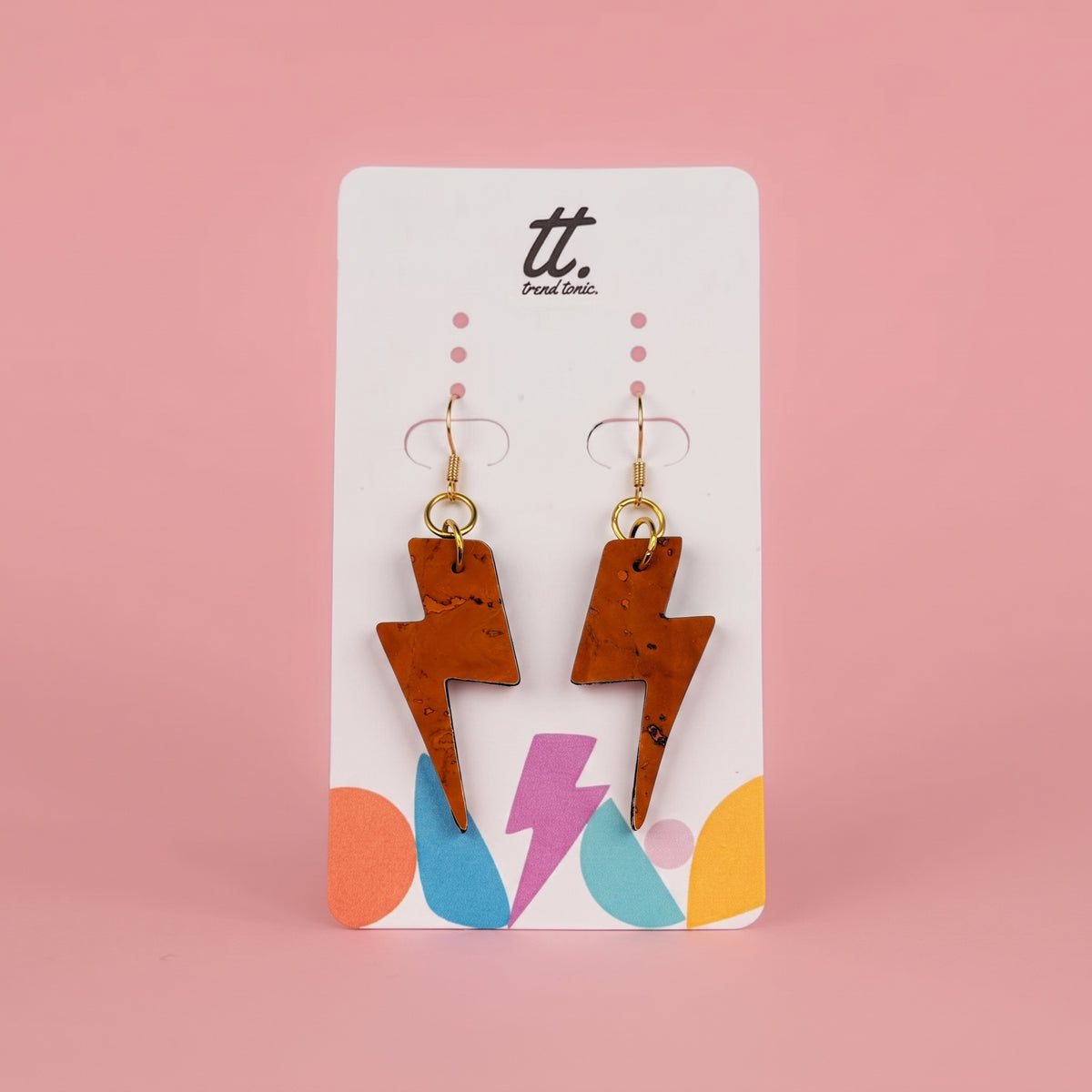 A pair of cork lightning bolt drop earrings with gold hooks, displayed on a white brand card with colorful accents against a solid pink background.