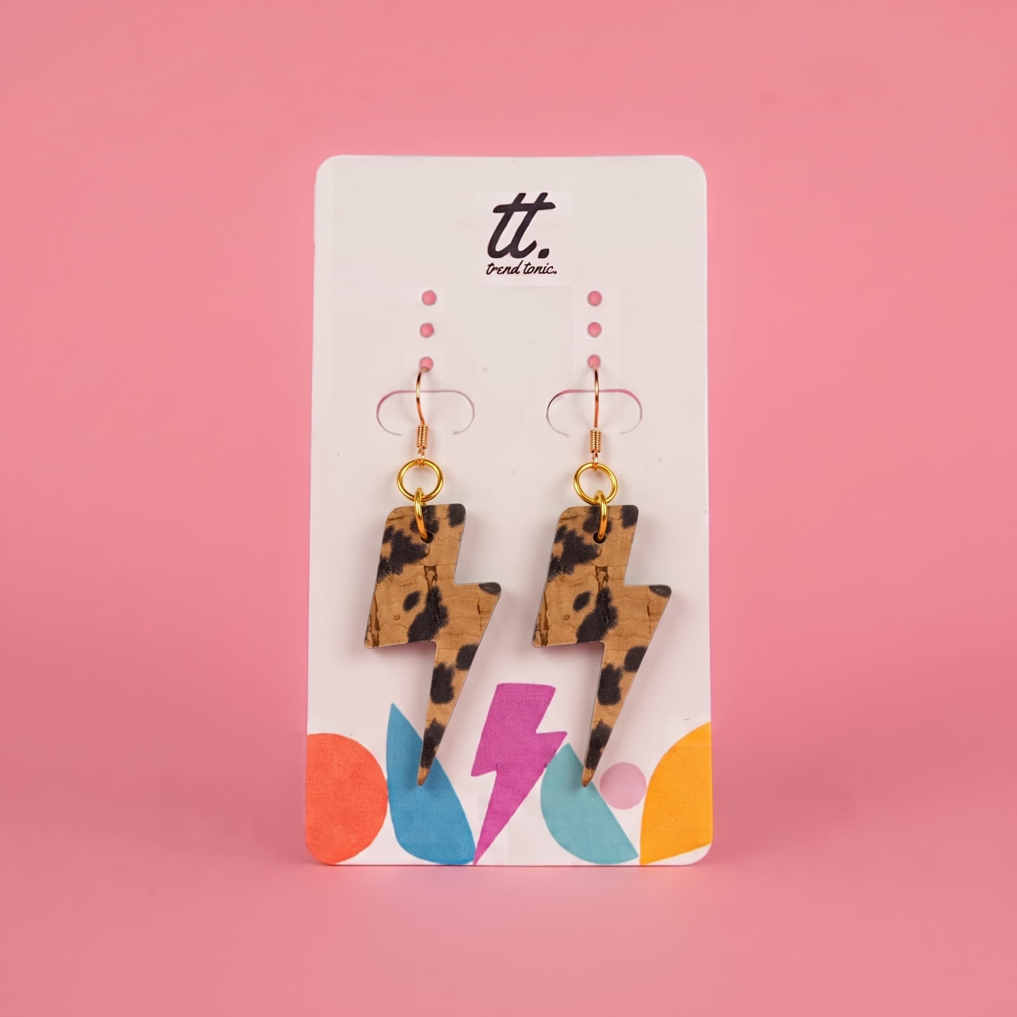 A pair of leopard-print lightning bolt drop earrings on a white display card with colorful graphic accents, set against a solid pink background. The brand logo 'tt.