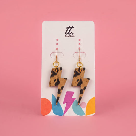 A pair of leopard-print lightning bolt drop earrings on a white display card with colorful graphic accents, set against a solid pink background. The brand logo 'tt.
