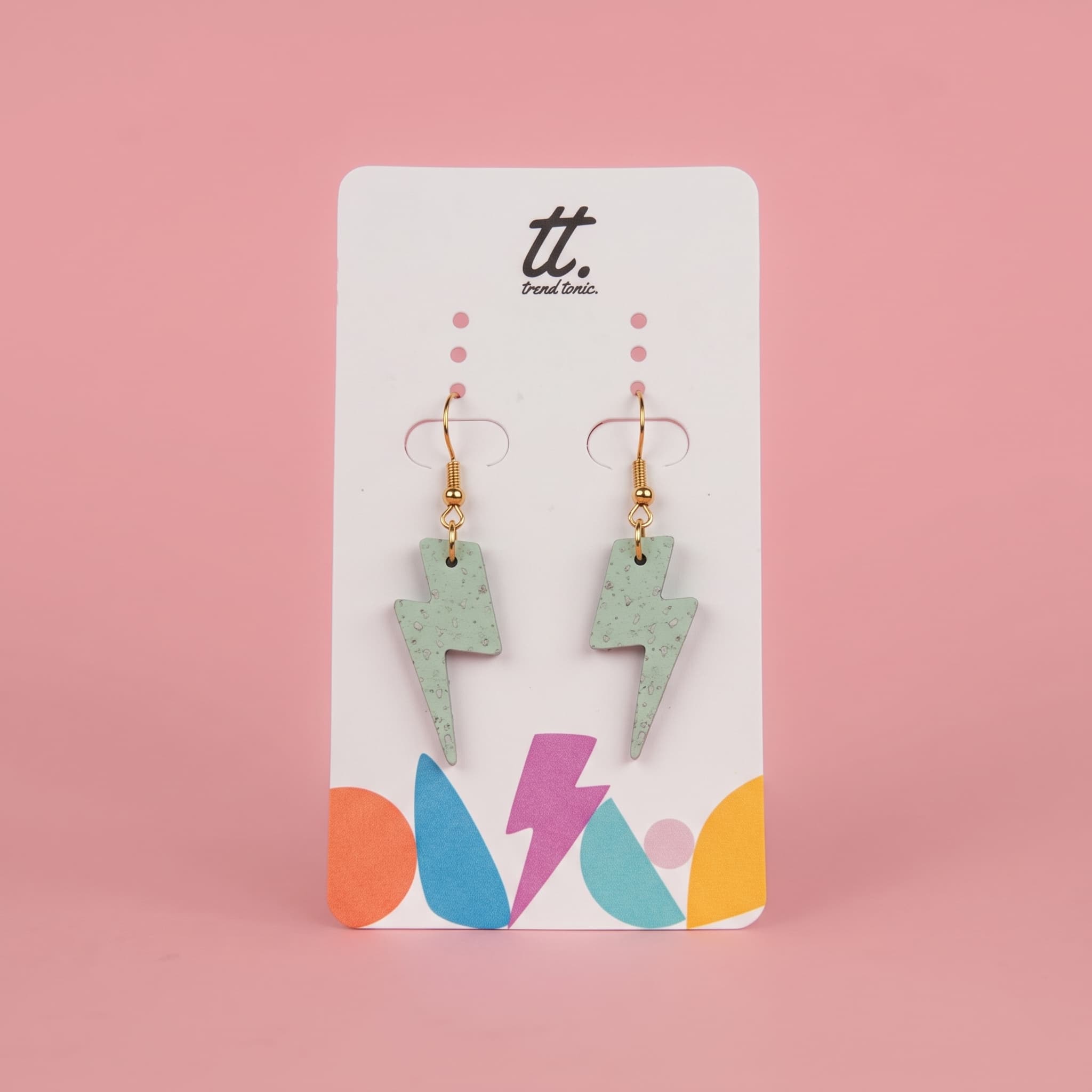 A pair of dangling lightning bolt earrings in a speckled mint green finish with gold hooks, displayed on a white card with colorful abstract shapes and the brand name 'tt. Paint Color.
