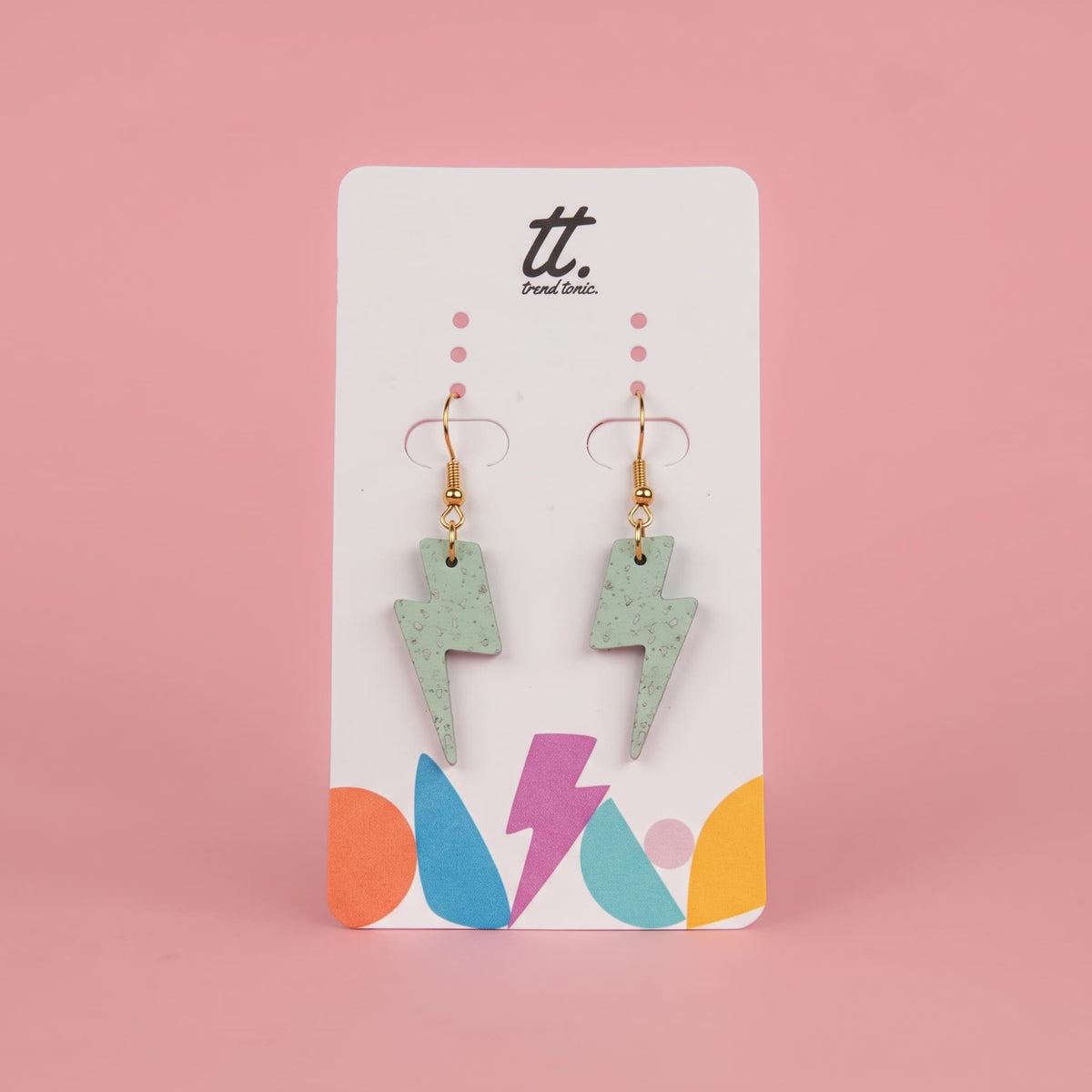 A pair of dangling lightning bolt earrings in a speckled mint green finish with gold hooks, displayed on a white card with colorful abstract shapes and the brand name 'tt. Paint Color.