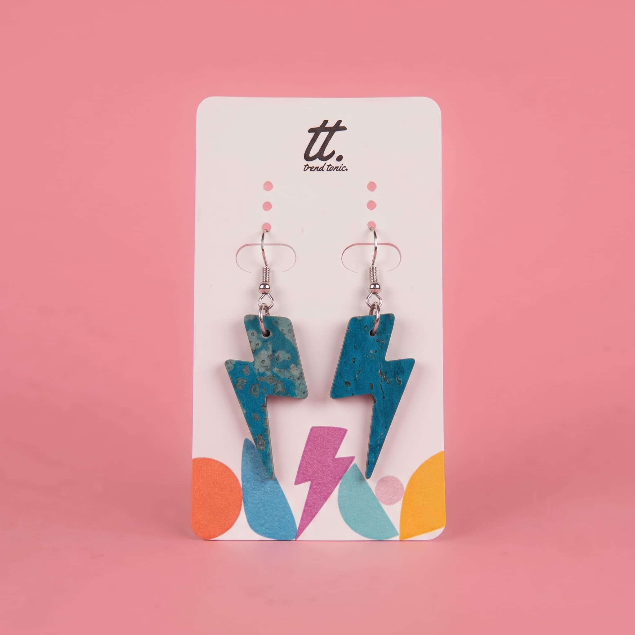 A pair of turquoise lightning bolt drop earrings with silver fishhook wires, displayed on a white brand card with colorful abstract shapes against a solid pink background.