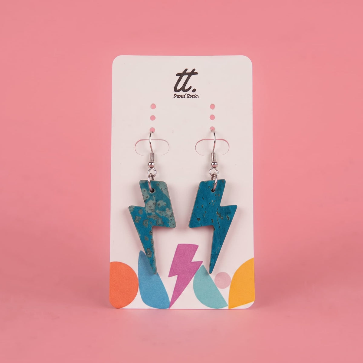 A pair of turquoise lightning bolt drop earrings with silver fishhook wires, displayed on a white brand card with colorful abstract shapes against a solid pink background.