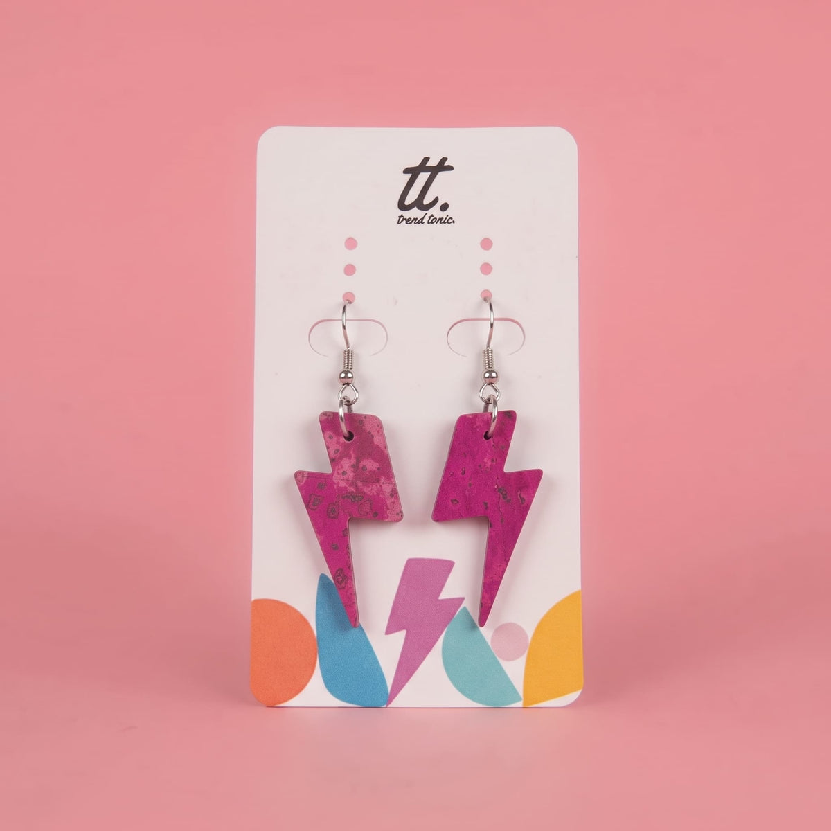 A pair of pink lightning bolt drop earrings on a white display card with colorful geometric shapes at the bottom, set against a solid pink background. The card features the brand logo 'tt.