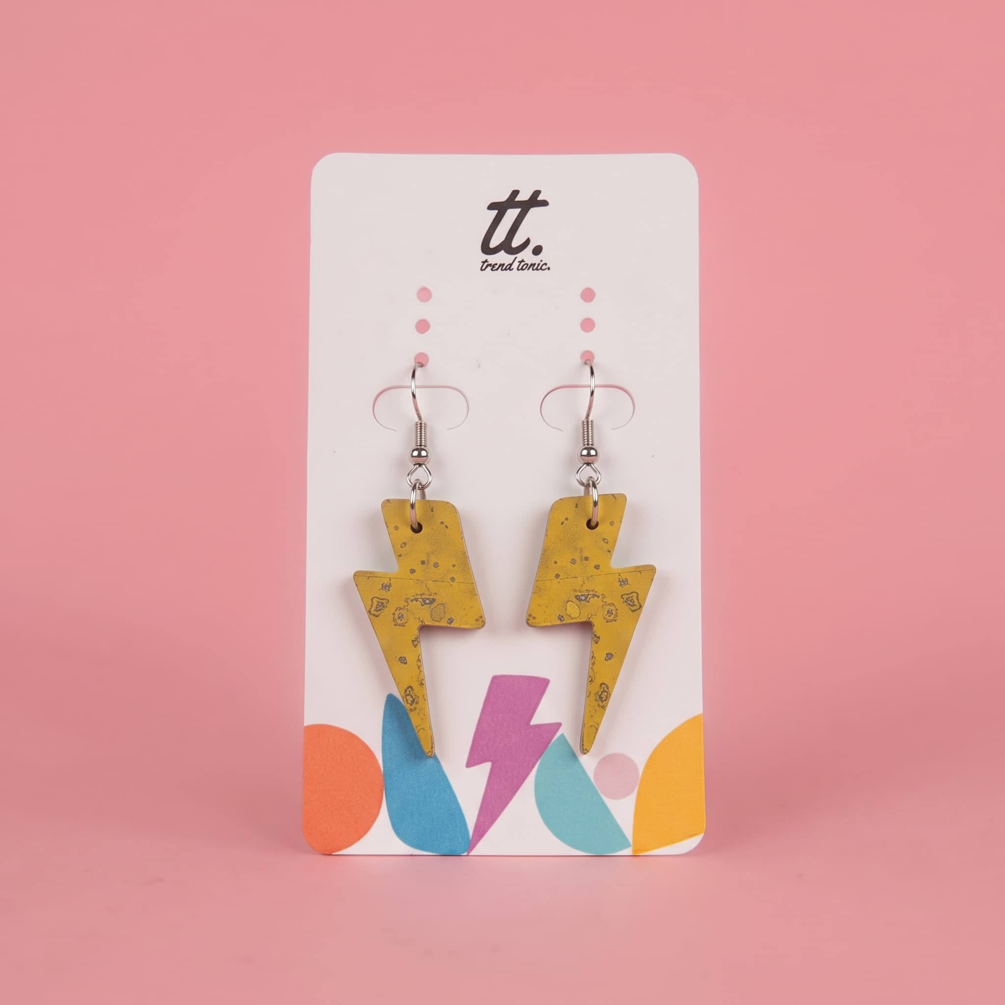A pair of yellow lightning bolt drop earrings with a subtle black speckled pattern, displayed on a white brand card with colorful geometric accents against a solid pink background.