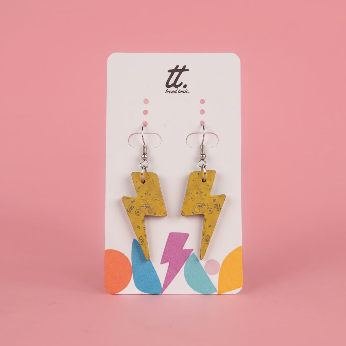 A pair of yellow lightning bolt drop earrings with a subtle black speckled pattern, displayed on a white brand card with colorful geometric accents against a solid pink background.
