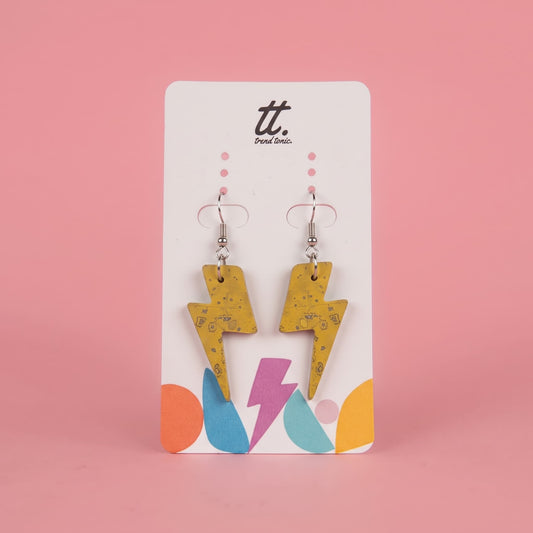 A pair of yellow lightning bolt drop earrings with a subtle black speckled pattern, displayed on a white brand card with colorful geometric accents against a solid pink background.